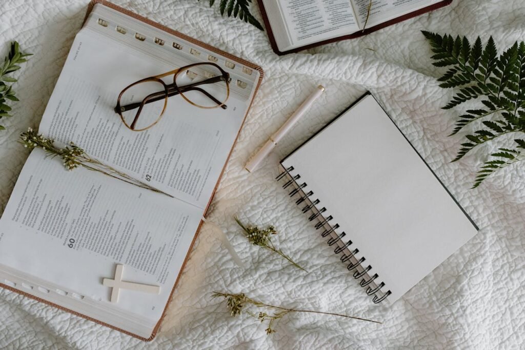 Open Bible, notebook, and eyeglasses on white bed with green leaves.