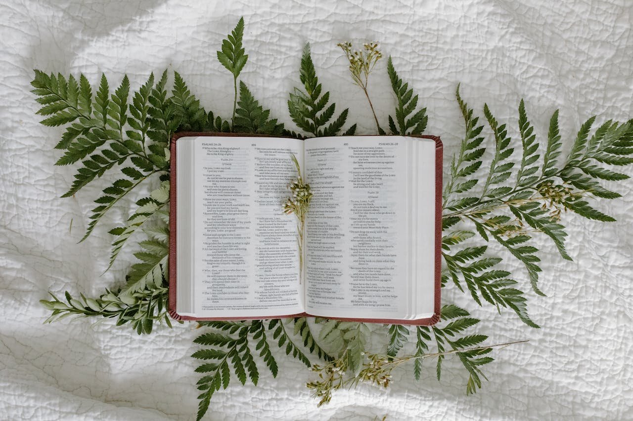 An open Bible displayed on a bedspread surrounded by fern leaves, symbolizing faith and nature.