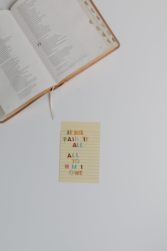 A flat lay of an open bible with a note stating 'Jesus paid it all, all to him I owe'.